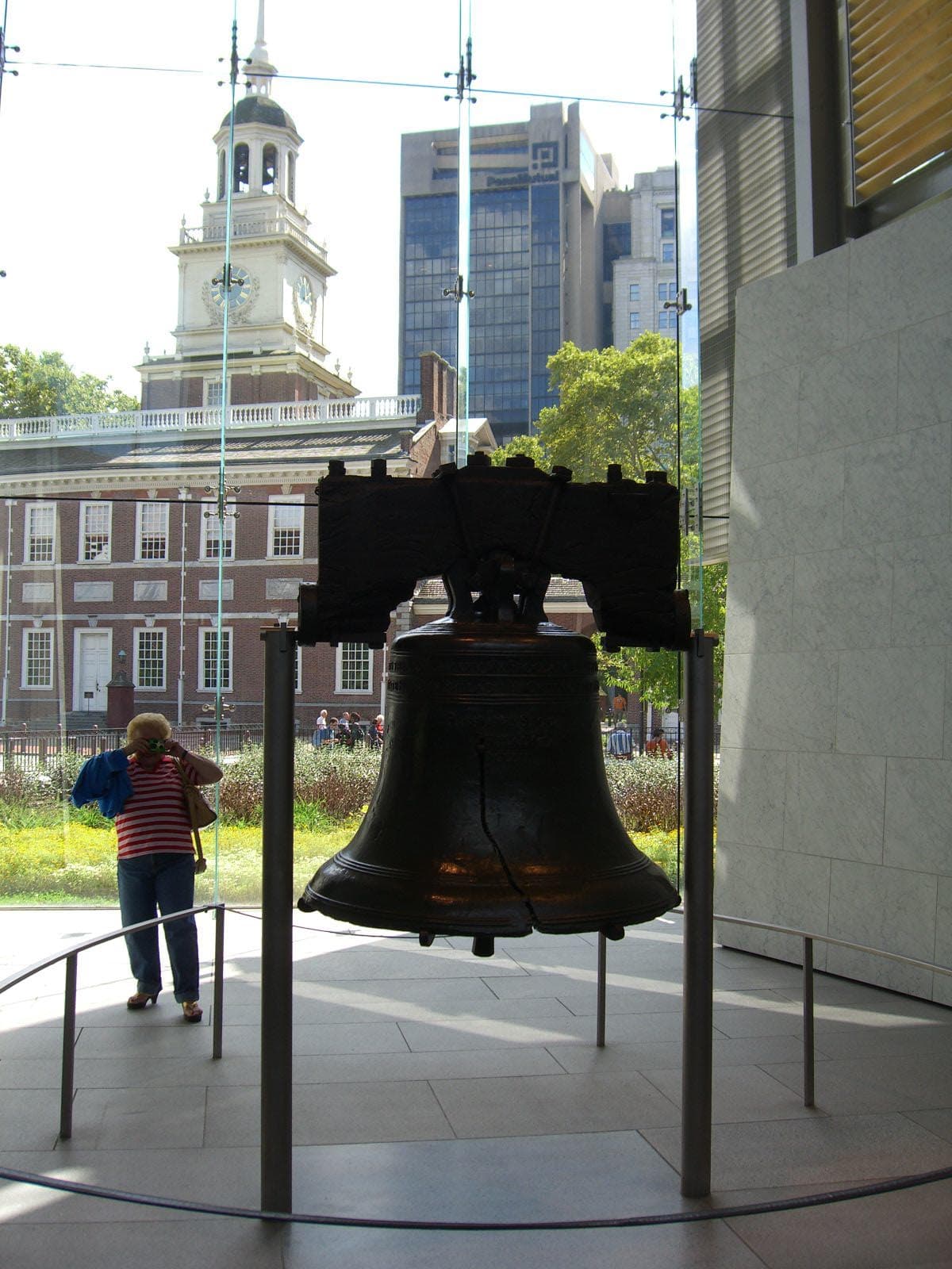 Hero photo: Luigi Novi (Nightscream), CC BY 3.0, via Wikimedia Commons. (The Liberty Bell in its glass chamber at the Liberty Bell Center, the famous crack facing the camera and Independence Hall — its home for two centuries — visible through the glass behind it.)
