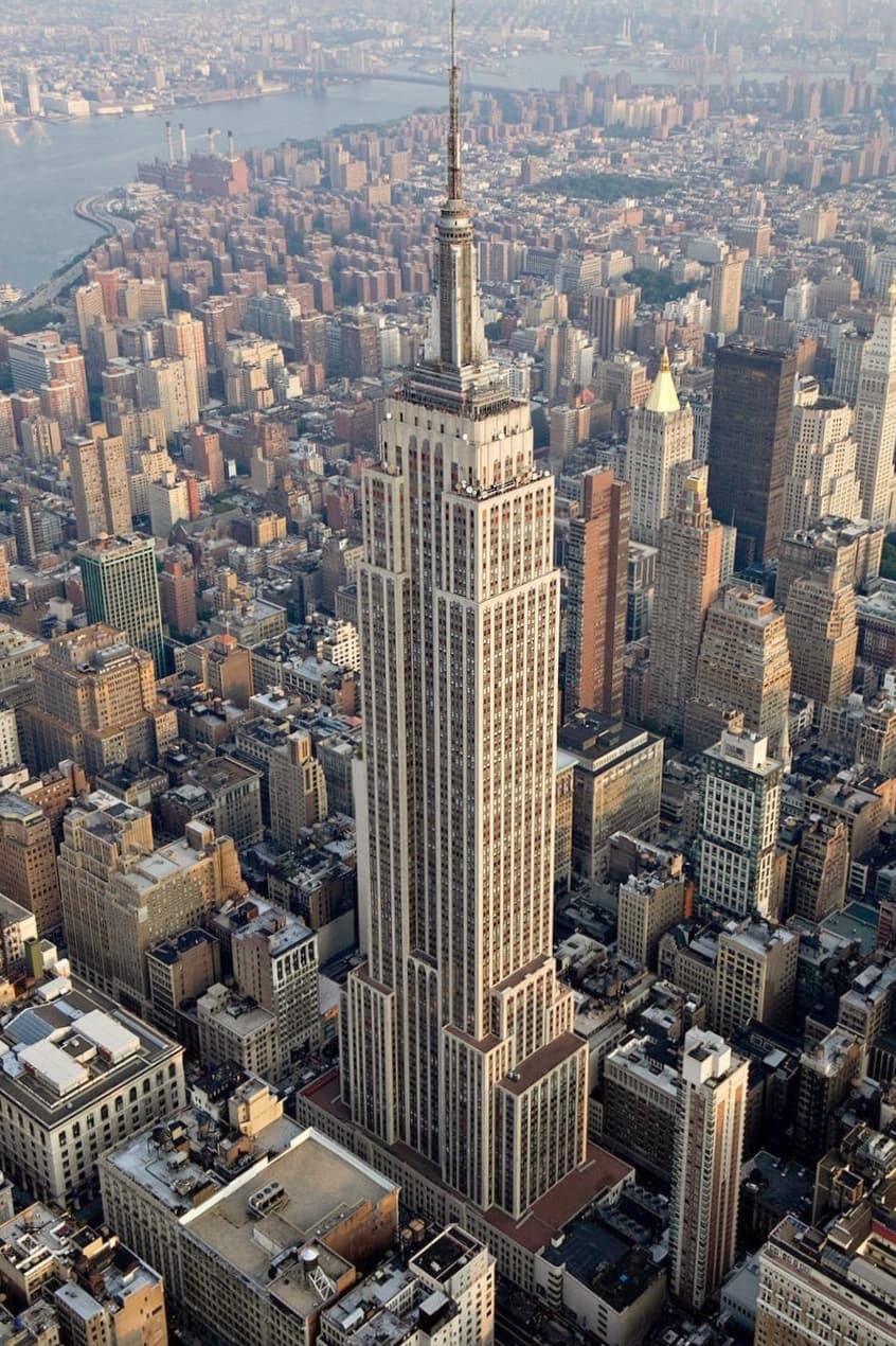 The Empire State Building from the air: the limestone shaft steps back floor by floor to the mooring mast, with the East River at upper left.