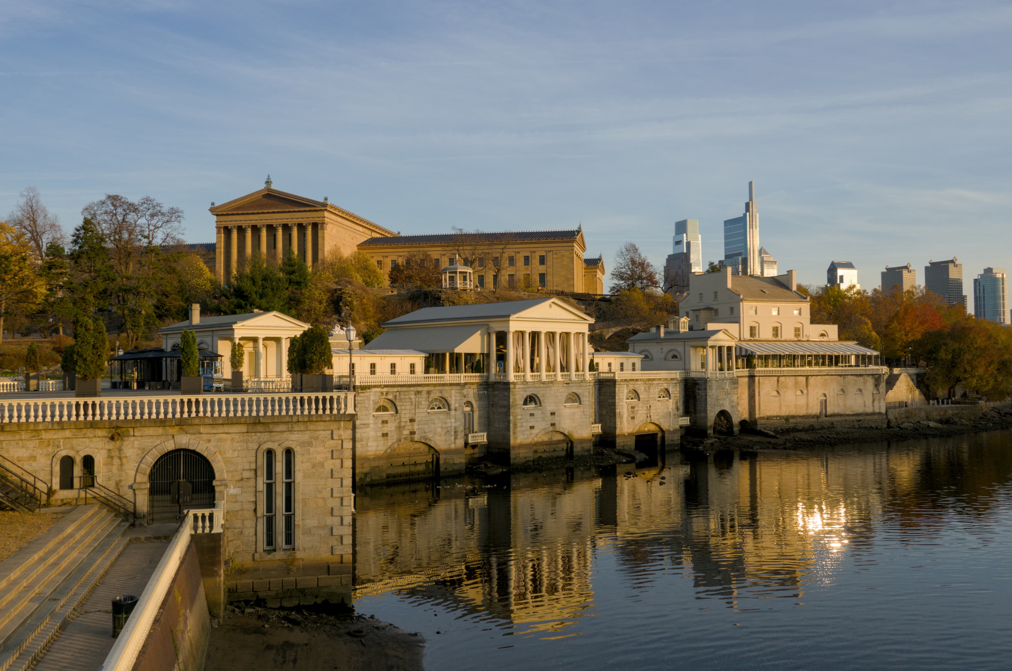 The Neoclassical mill houses of the Fairmount Water Works reflected in the Schuylkill River at golden hour, with the Philadelphia Museum of Art on Faire Mount above and the Center City skyline behind — the reservoir Graff pumped to stood where the museum now is.