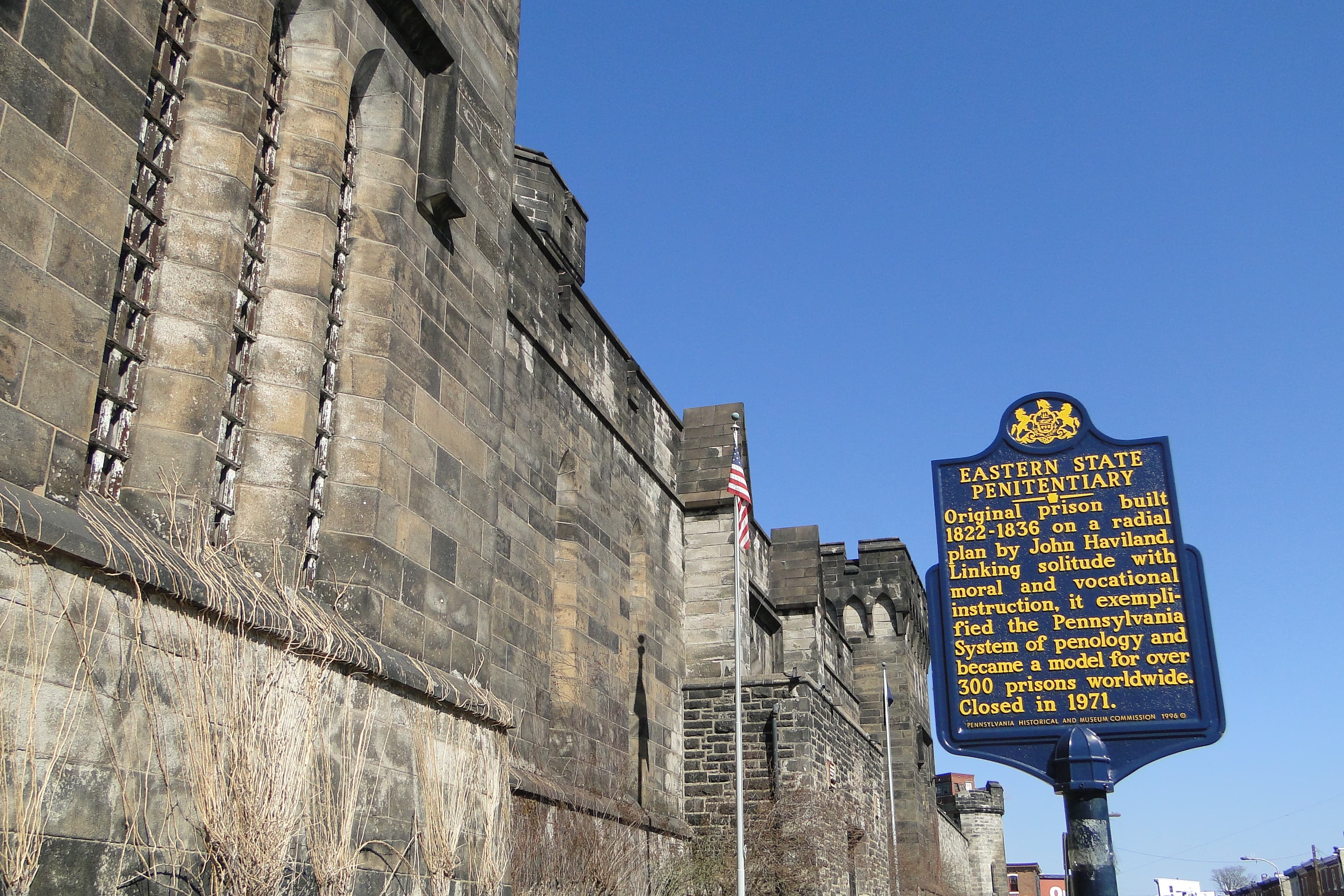 The stone front wall and towers of Eastern State Penitentiary on Fairmount Avenue, with the blue-and-gold Pennsylvania historical marker for the prison in the right foreground — this is the front wall, not the prison entrance or interior.